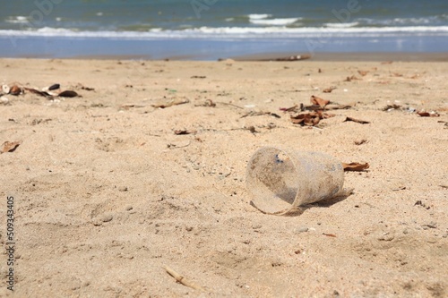 Garbage, plastic glass, on the beach with shells on sand in summer morning. Environment and nature concept.