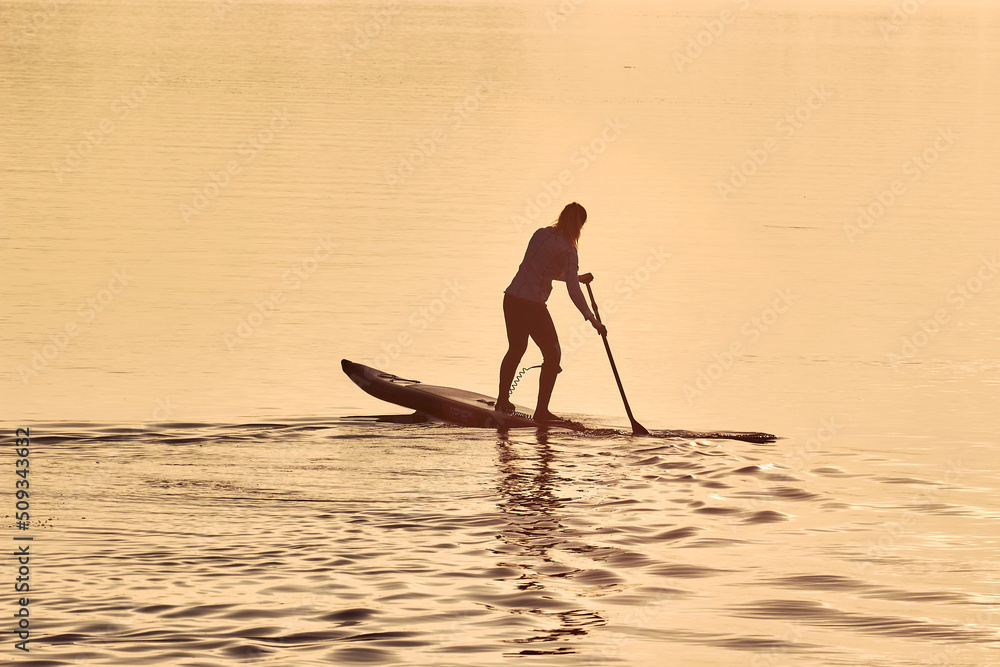Silhouette of woman paddle on stand up paddle board (SUP) on quiet winter or autumn river at sunset. Water sport and meditation on the water