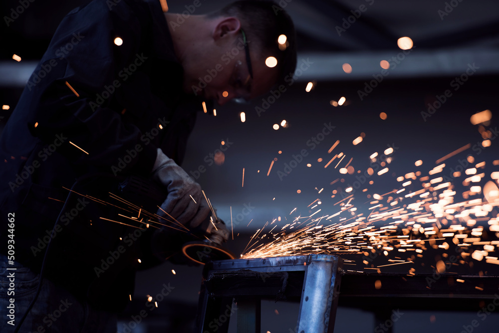 Heavy Industry Engineering Factory Interior with Industrial Worker Using Angle Grinder and Cutting a Metal Tube. Contractor in Safety Uniform and Hard Hat Manufacturing Metal Structures. Heavy Industry Engineering Factory Interior with Industrial Worker Using Angle Grinder and Cutting a Metal Tube. Contractor in Safety Uniform and Hard Hat Manufacturing Metal Structures.