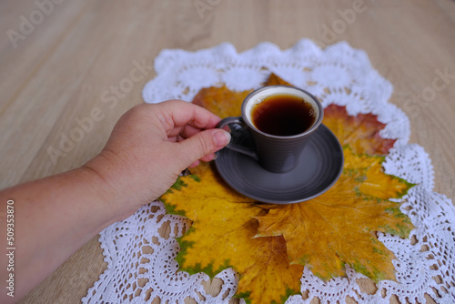 close-up of female hand holding a gray cup of coffee, yellow wedge leaves picturesquely lie on a lacy white napkin, time to drink tea and coffee, effect of caffeine on health, concept of autumn mood photo