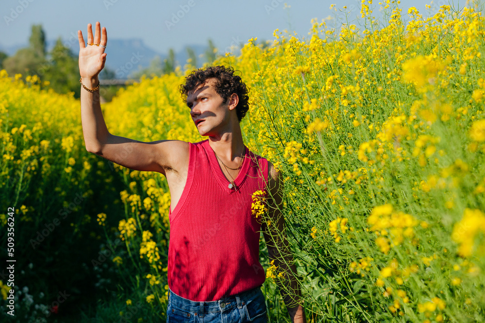 Young homosexual man with curly hair and raised hand, covers his face from the sunlight, standing at yellow oilseed rapes, flower field at spring. Young homosexual man with curly hair and raised hand, covers his face from the sunlight, standing at yellow oilseed rapes, flower field at spring.