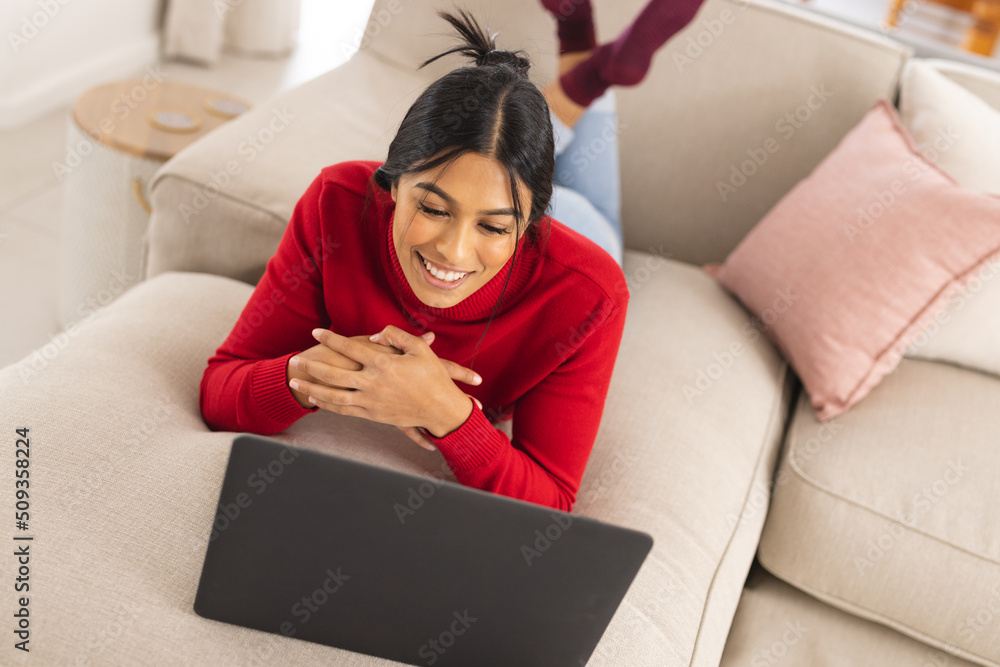 Happy biracial young woman video calling through laptop while lying on ...