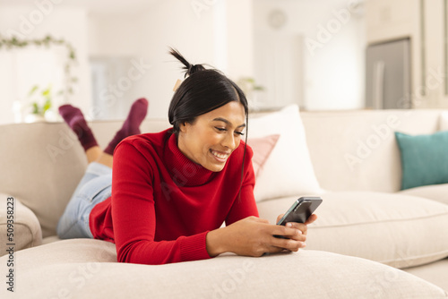 Smiling biracial young woman using smart phone while lying on sofa at home