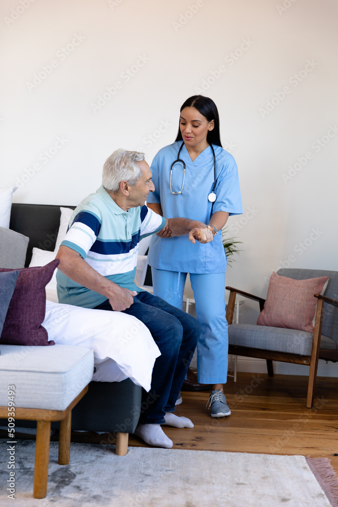 Biracial female physiotherapist assisting caucasian senior man in getting up from bed at home