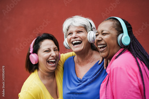 Happy mature female friends having fun together while listeing music with wireless headphone - Multiethnic women enjoy day outdoor