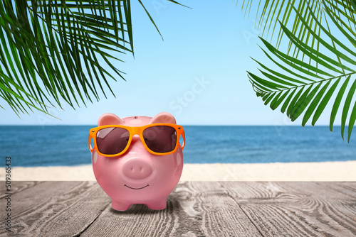 Saving money for summer vacation. Piggy bank with sunglasses on wooden surface near sandy beach and sea © New Africa