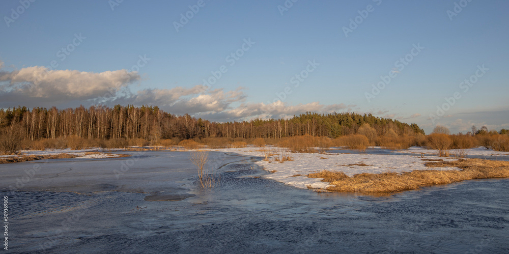 March sunny evening by the river. Blue sky over the horizon. A picturesque landscape, early spring, a river with snow-covered banks, dry grass and bushes. The first thaws, the snow is melting March sunny evening by the river. Blue sky over the horizon. A picturesque landscape, early spring, a river with snow-covered banks, dry grass and bushes. The first thaws, the snow is melting