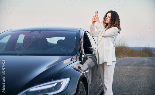 Beautiful curly woman with cute smile standing near her black luxury electric car on road, and taking picture at phone on the backdrop of grey sky. © anatoliy_gleb