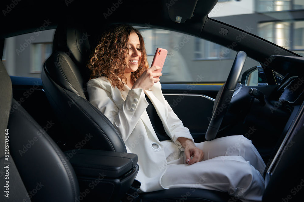 View from inside automobile. Young girl looking at smartphone while in the driver's seat in electric car with vehicle autopilot system. Likable woman using cellphone in car interior and cute smiling.
