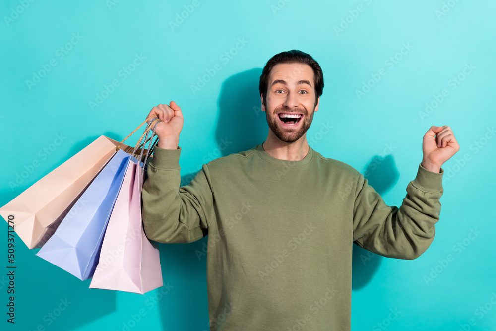 Photo of young excited guy rejoice victory fists hands hold shopping bags store isolated over turquoise color background Photo of young excited guy rejoice victory fists hands hold shopping bags store isolated over turquoise color background