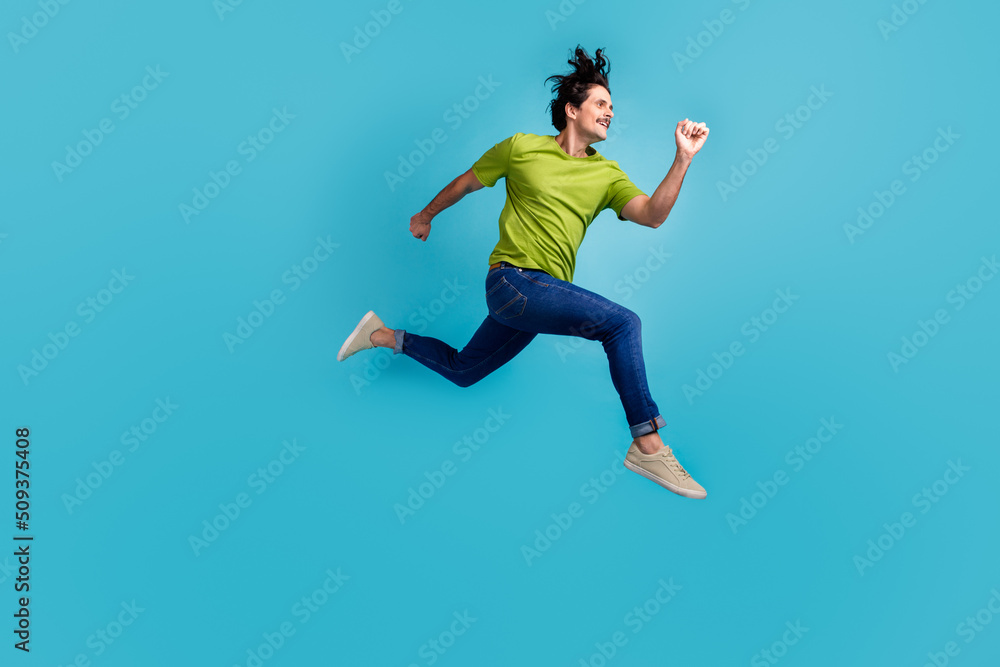 Full length photo of positive excited man wear green t-shirt hurrying jumping high isolated blue color background Full length photo of positive excited man wear green t-shirt hurrying jumping high isolated blue color background