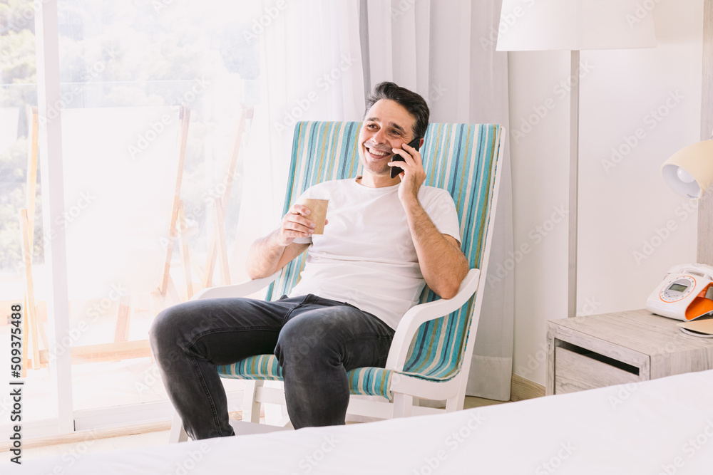 Young man sitting in a rocking chair in his bedroom, talking on the mobile and having a coffee, with the light coming through the window. Concept of working, vacation, connection and smartphone. Young man sitting in a rocking chair in his bedroom, talking on the mobile and having a coffee, with the light coming through the window. Concept of working, vacation, connection and smartphone.