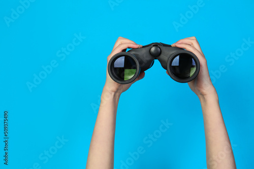 Woman holding modern binoculars on light blue background, closeup. Space for text photo
