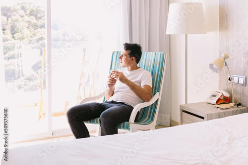 Young man sitting in a rocking chair in his bedroom, having a drink with the light coming through the window. Vacation, relaxation, rest, breakfast, coffee, tea and hotel concept.