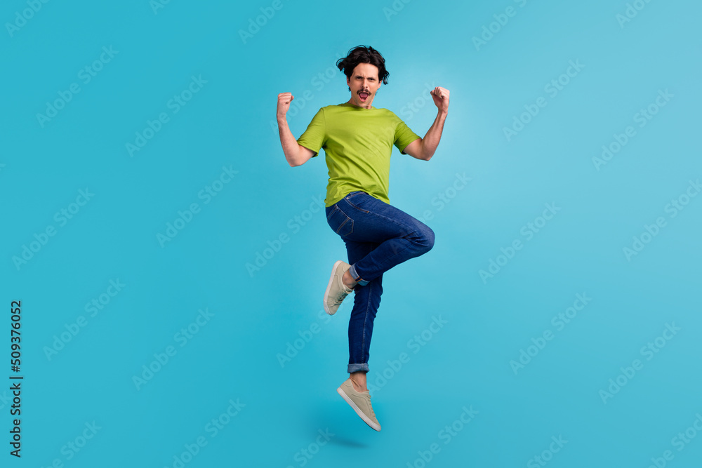 Full length photo of funky lucky guy dressed green t-shirt jumping high rising fists isolated blue color background