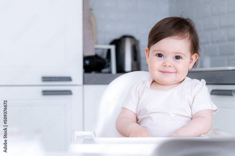 Portrait cute baby girl on kitchen sitting on high chair, smiling child happy to eat first solid food.