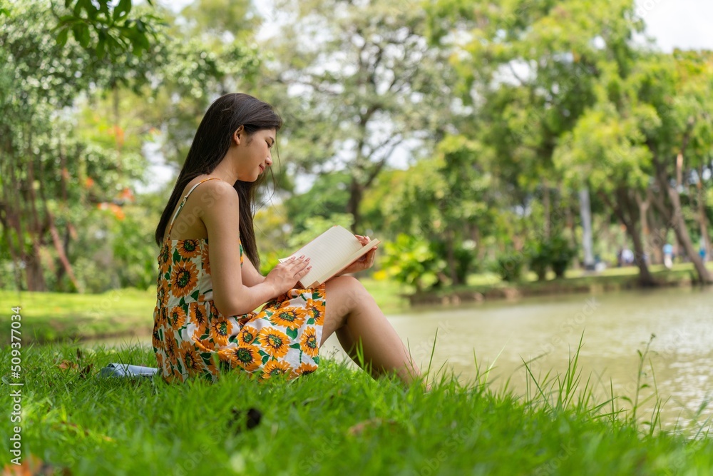 Young beautiful asian woman in sunflower summer dress sitting on the grass reading and mesmerizing a book and smiling happily while enjoying her free time in a public park Young beautiful asian woman in sunflower summer dress sitting on the grass reading and mesmerizing a book and smiling happily while enjoying her free time in a public park