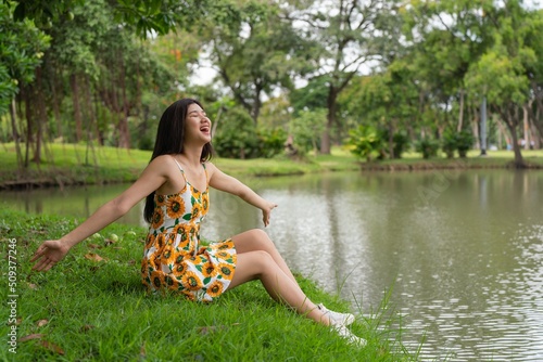 Portrait photo of a young beautiful asian female lady in sunflower summer dress pattern sitting on the grass embracing the breeze wind and soft sunlight relaxingly and chillingly in a public park photo