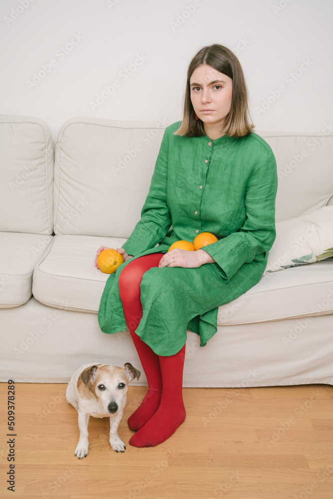 Green dress and red tights. creative composition of color contrasts. indifferent mood. dog sitting on the floor. Girl holding Oranges sitting on beige sofa. Concept idea creative image. focus on girl Green dress and red tights. creative composition of color contrasts. indifferent mood. dog sitting on the floor. Girl holding Oranges sitting on beige sofa. Concept idea creative image. focus on girl