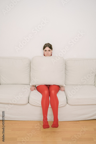 apathy and depression. Young beautiful girl in red tights sits on beige sofa and hides behind cushion. looking indifferent and unemotional. Vertical creative composition
