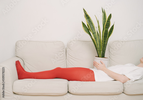 girl in red tights lies on a beige sofa with houseplant on her stomach. waiting is tired. Artistic creative conceptual  composition. Horizontal. restrained emotions in  appointment with psychotherapis