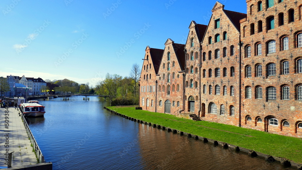 5 Salzspeicher in Lübeck entlang von Fluss Trave mit  Rasen und Schiffsanlegestelle unter blauem Himmel