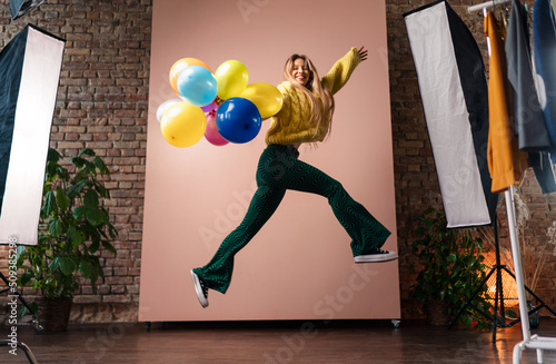 Fashion studio portrait of a happy young woman with balloons jumping, backstage of photoshooting .