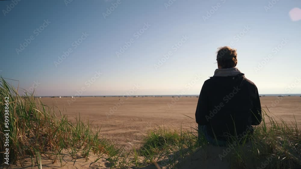 Lonely man sitting on sand dune at a beach in denmark watching the horizon