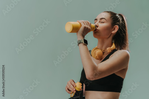 Young sporty woman in sportswear with wireless headset drinking from bottle over green background, copy space