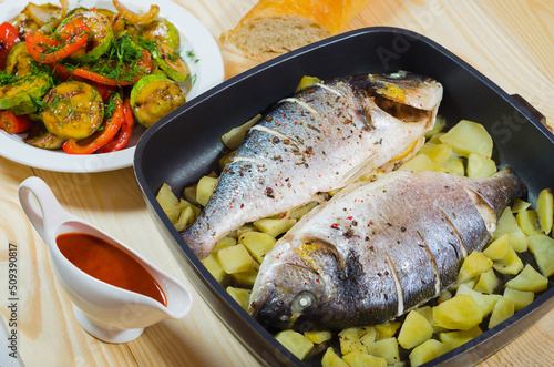 Two baked fish and potato in pan at wooden table with sauce and vegetables