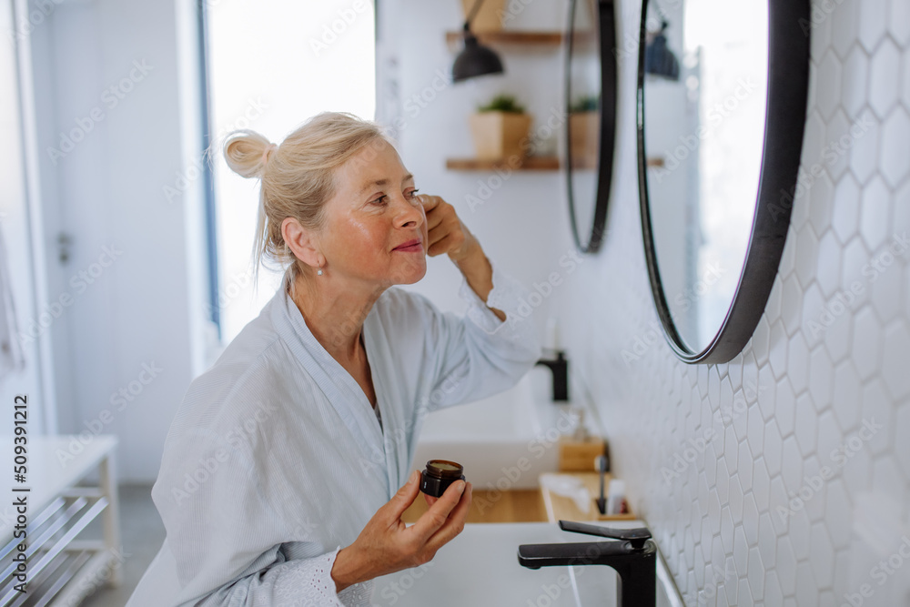 Obraz premium Beautiful senior woman in bathrobe applying natural face cream in bathroom, skin care and morning routine concept.
