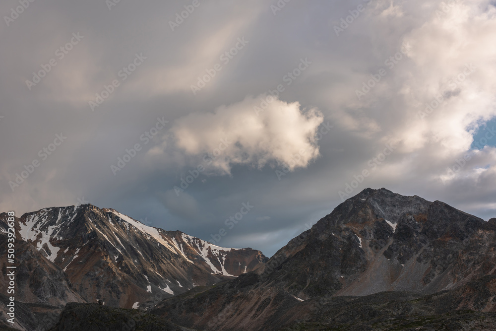 Obraz premium Dramatic mountain landscape with high mountain range with sharp rocky pinnacle under clouds of sunset color in gloomy sky. Dark atmospheric scenery with large mountains with snow at changeable weather