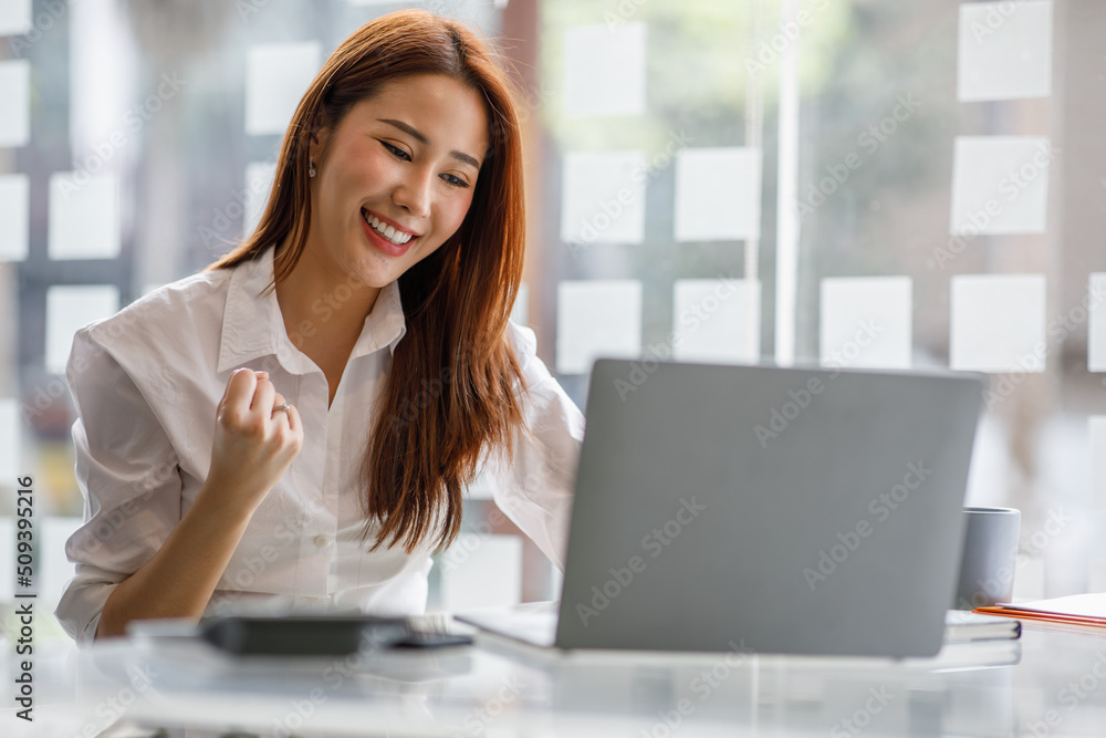 Portrait of happy young Asian woman celebrating success with arms up in front of a laptop at office