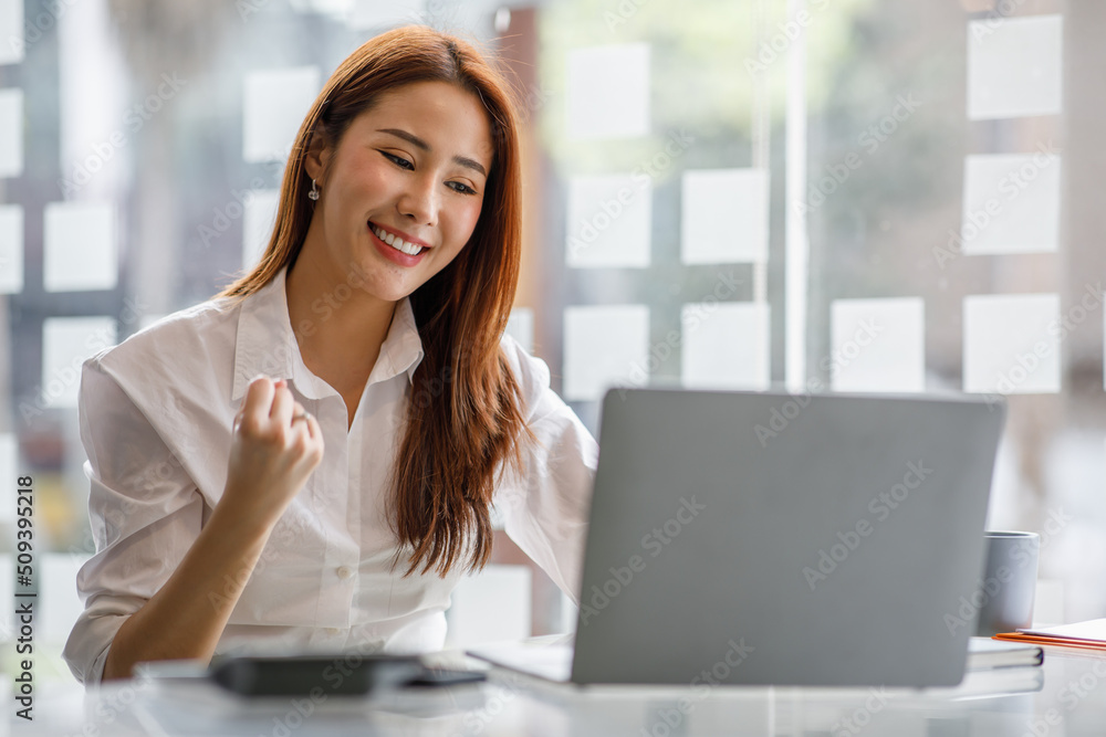 Portrait of happy young Asian woman celebrating success with arms up in front of a laptop at office Portrait of happy young Asian woman celebrating success with arms up in front of a laptop at office