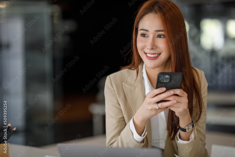 Photo of the adorable young lady working on laptop computer and holding smartphone at office indoors, Asian business woman.