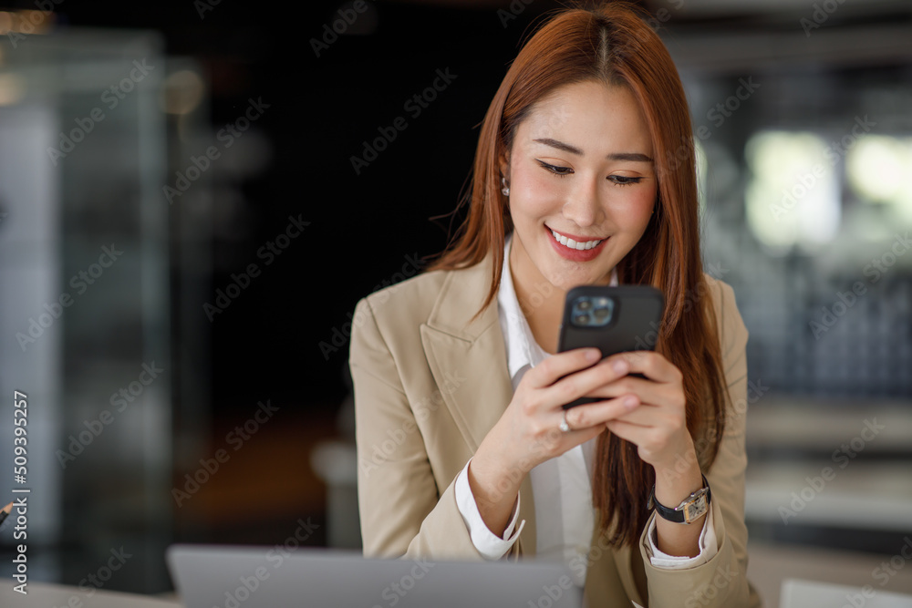 Photo of the adorable young lady working on laptop computer and holding smartphone at office indoors, Asian business woman.
