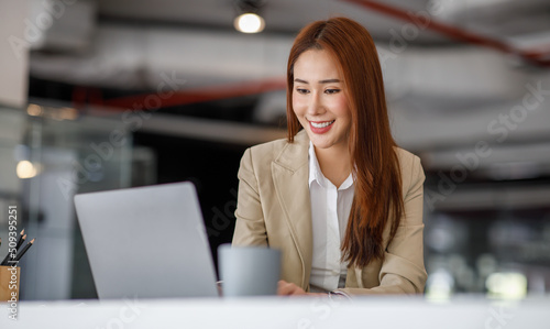Beautiful young Asian businesswoman in casual clothing using a laptop and smiling while working indoors