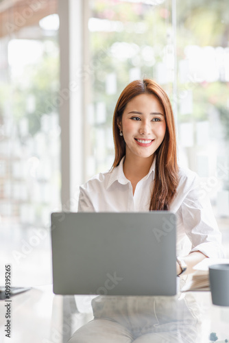 Photo of the adorable young lady working on laptop computer and holding smartphone at office indoors, Asian business woman.