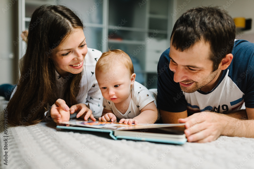 Happy family, parenthood and people concept - mother, father with baby lying in bed at home. Portrait of young smiling family with son reading book. Happy family, parenthood and people concept - mother, father with baby lying in bed at home. Portrait of young smiling family with son reading book.