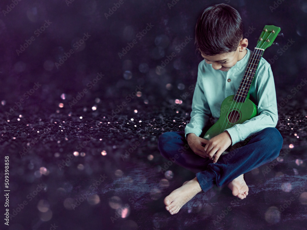 Naklejka premium latino boy sitting on the floor with a ukulele in his arms laughing and looking down. Studio photo with black background and copy space. Concept of learning to play an instrument in childhood