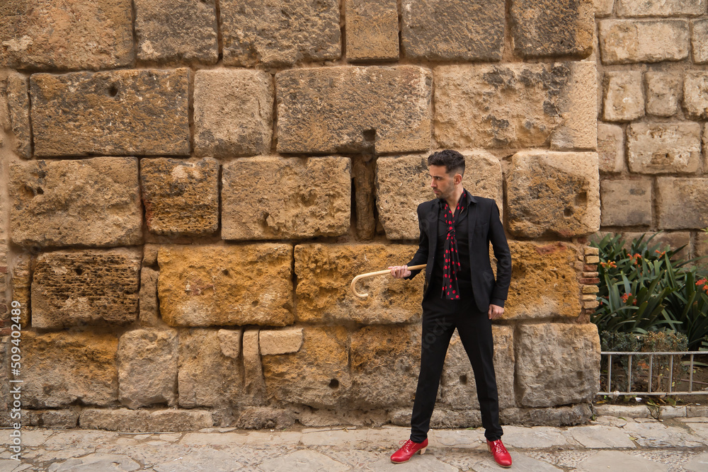 Young Spanish man in black shirt, jacket and pants, with black scarf with red polka dots, red shoes and walking stick posing next to a stone wall. Flamenco concept, art, dance, culture, tradition. Young Spanish man in black shirt, jacket and pants, with black scarf with red polka dots, red shoes and walking stick posing next to a stone wall. Flamenco concept, art, dance, culture, tradition.