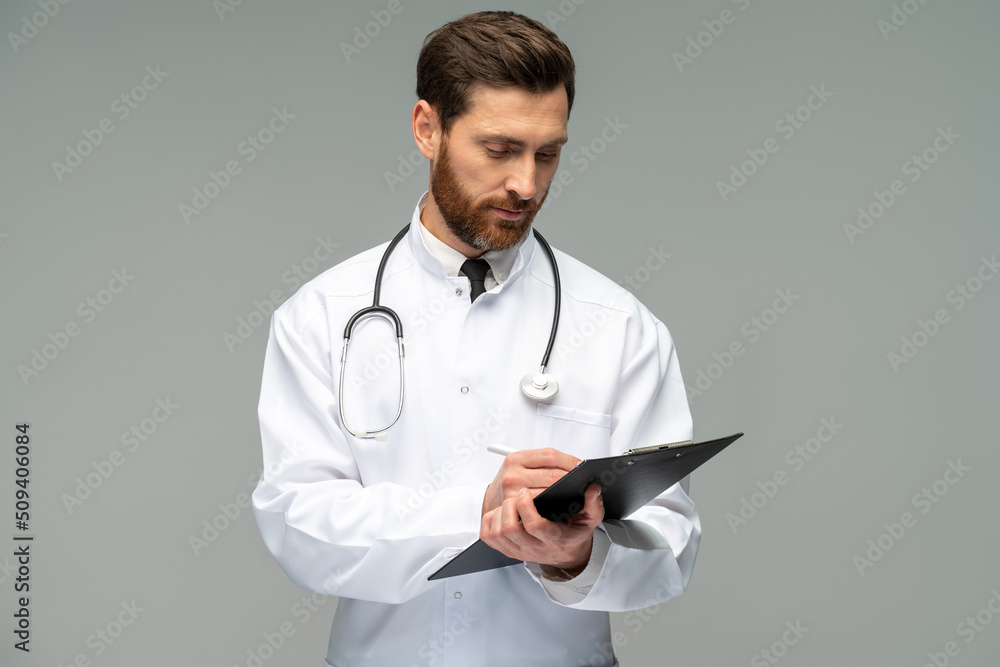 Happy caucasian doctor with stethoscope holding clipboard and writing something with concentrated face while standing isolated on grey background Happy caucasian doctor with stethoscope holding clipboard and writing something with concentrated face while standing isolated on grey background