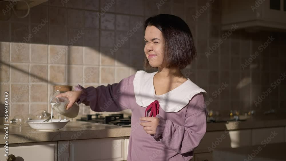 Adult little woman pouring milk in morning cereals smiling looking out the window. Portrait of confident positive Caucasian little person enjoying breakfast at home indoors Adult little woman pouring milk in morning cereals smiling looking out the window. Portrait of confident positive Caucasian little person enjoying breakfast at home indoors