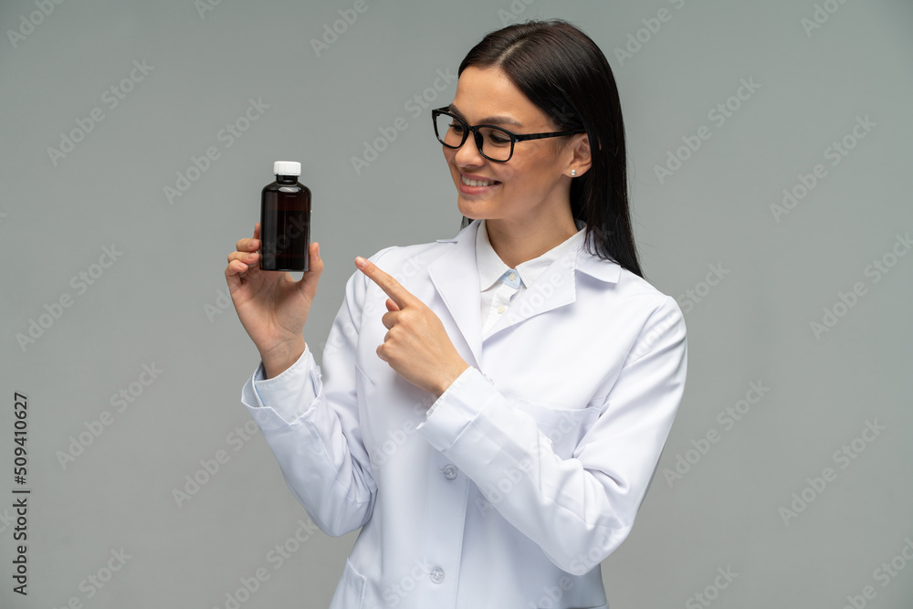 Waist up portrait view of the female doctor wearing white coat holding bottle with mixture and looking on it over grey background Waist up portrait view of the female doctor wearing white coat holding bottle with mixture and looking on it over grey background