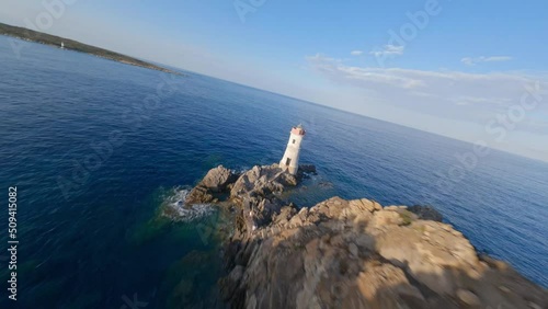 FPV video, view from above, stunning aerial view from an FPV drone flying at high speed over a rocky coastline with a lighthouse illuminated during a dramatic sunset. Faro di Capo Ferro, Sardinia.