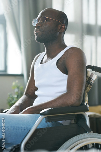 African American man in eyeglasses with disability sitting in wheelchair and looking away with pensive expression