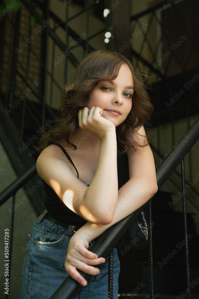 Fototapeta premium Pretty teen girl with curly hair smiles and looks to the camera. Portrait of beautiful young teenager little girl with nice hairstyle. Close-up, selective focus