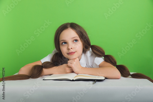 Cute little girl schoolgirl looks up. Child with books sits lost in thought on a green background.