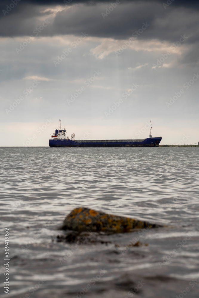 Cargo ship in focus. Rough stone rocks in foreground out of focus. Danger of the ocean. World goods shipping industry. Export and import business. Dark and moody sky and water surface. Cargo ship in focus. Rough stone rocks in foreground out of focus. Danger of the ocean. World goods shipping industry. Export and import business. Dark and moody sky and water surface.