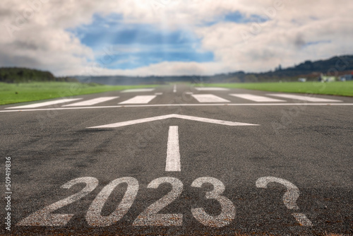 White arrow on a small asphalt air plane runway and sign 2023 with question mark. Cloudy sky with small blue opening in the middle. Sun rays and flare. Small airport on the right. Selective focus